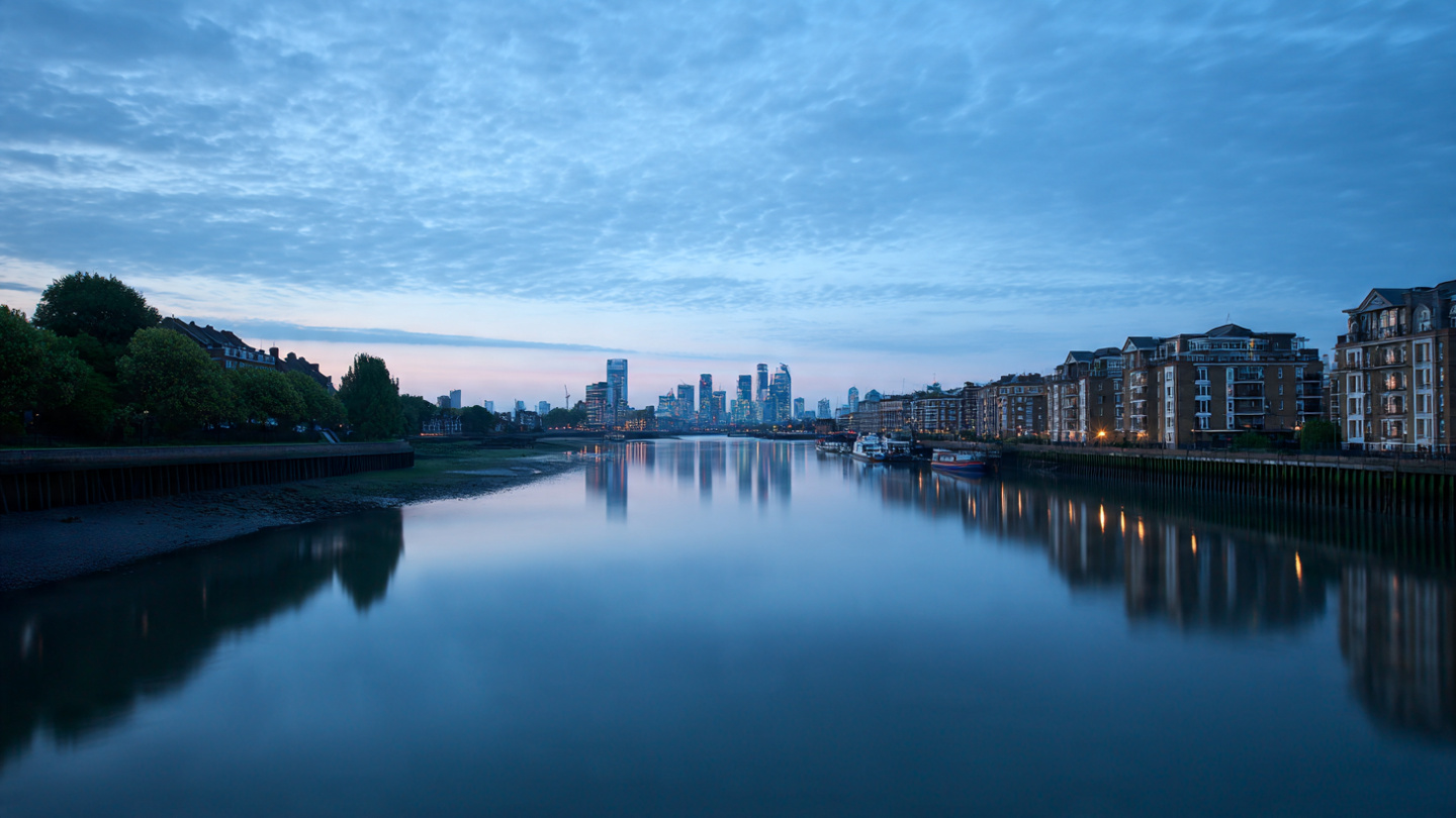 Hidden viewpoints in London along a peaceful riverside at dawn, showing a calm reflective skyline and waterfront buildings, capturing a serene secret viewpoint and quiet scenic river view of the London skyline.