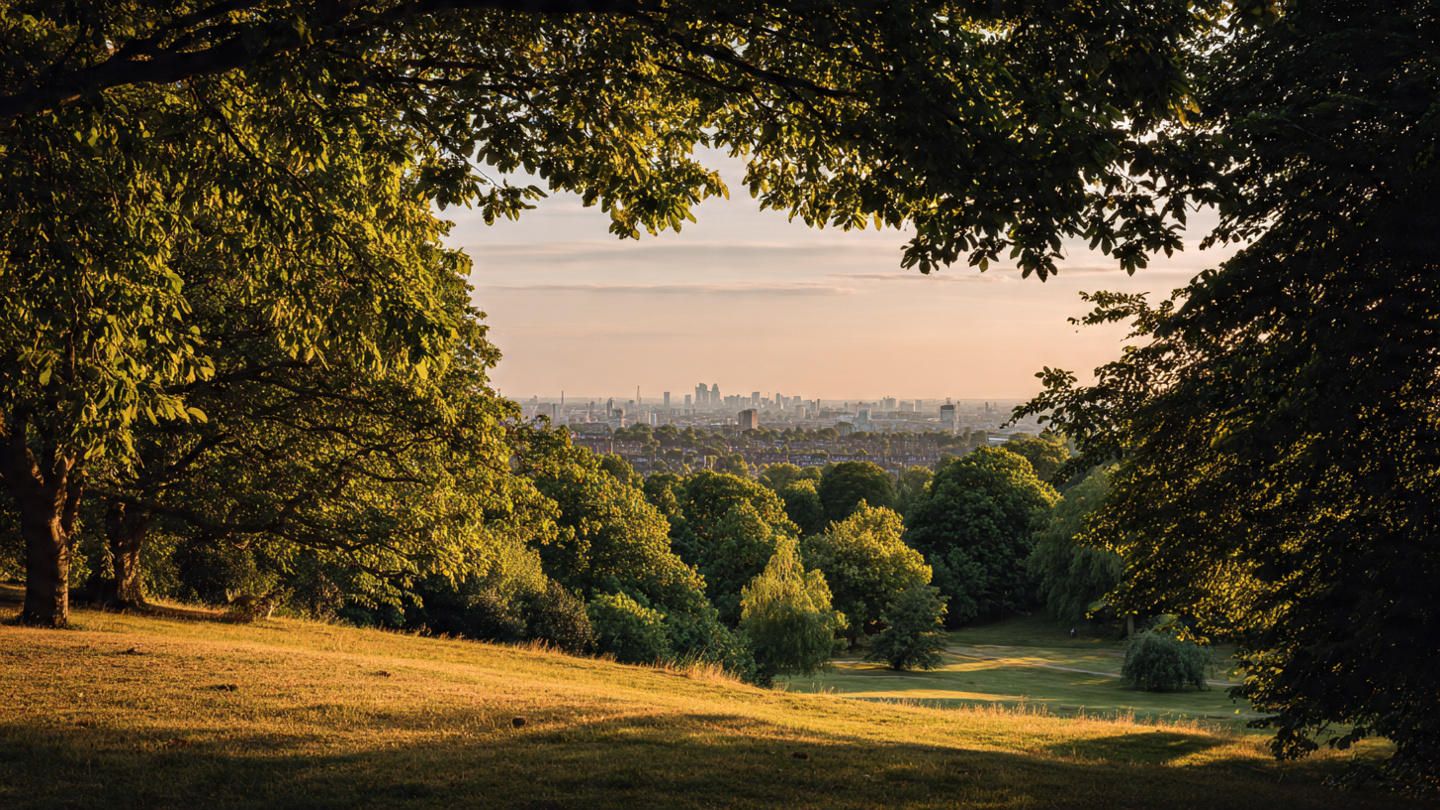 Hidden viewpoints in London from a peaceful hillside park at sunset, with trees framing a distant skyline and soft golden light, capturing a scenic secret viewpoint and quiet park outlook over the London city skyline.