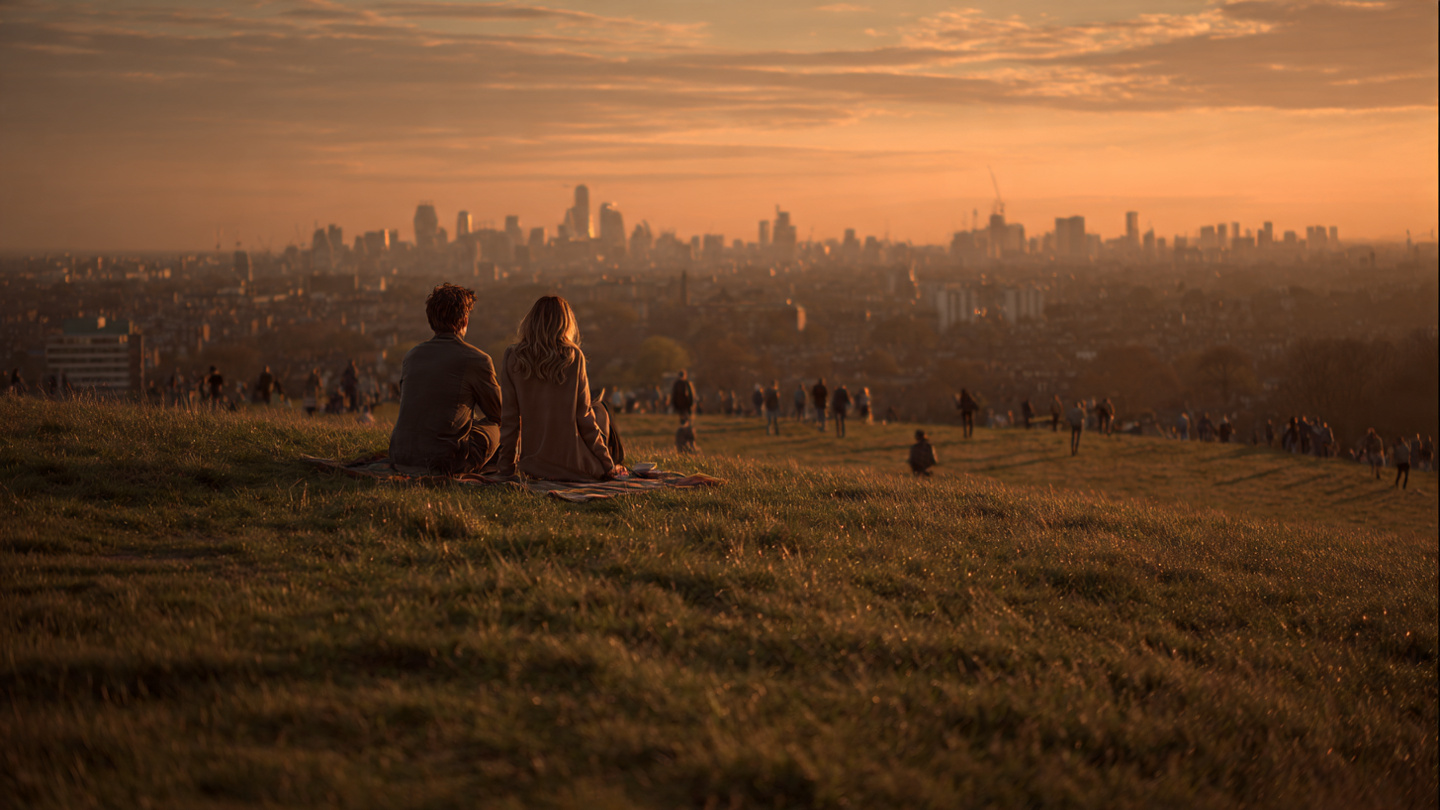 romantic walks in london at sunset from Primrose Hill with a couple sitting on the grass overlooking the golden skyline