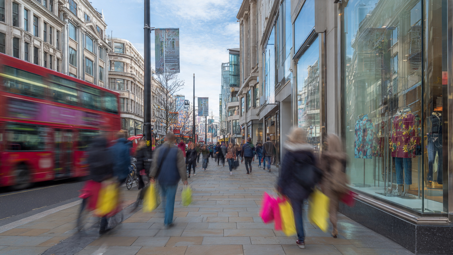 Busy daytime scene on one of the best shopping streets in London with shoppers walking past glass storefronts and a red double-decker bus in motion